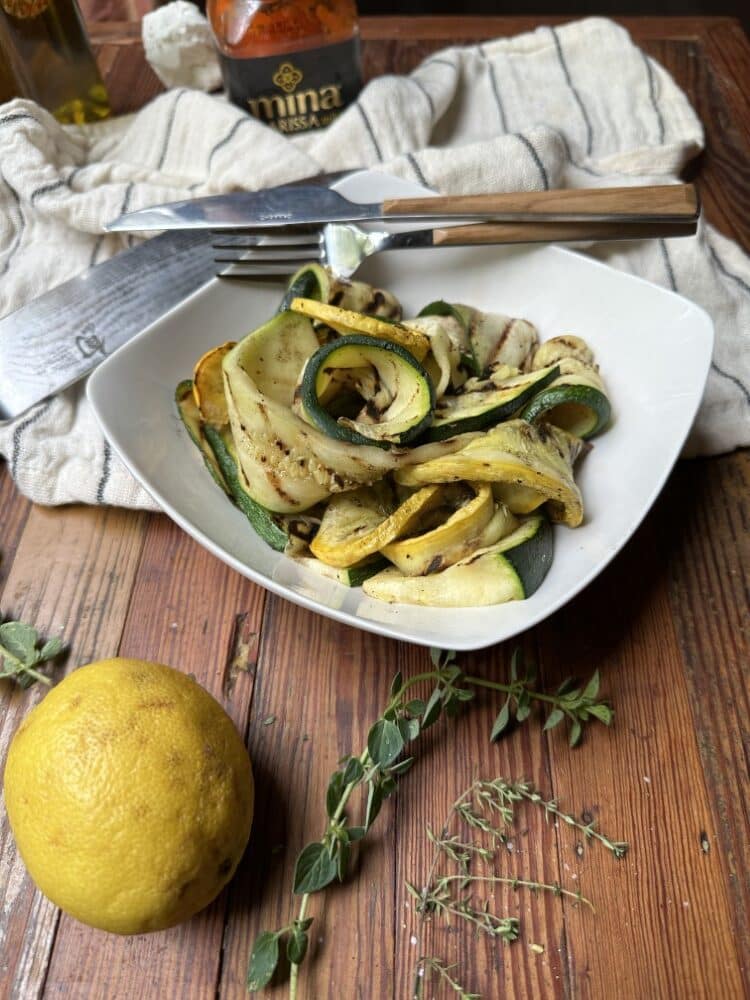 serving of grilled zucchini salad in white, shallow square bowl with fork and knife, lemon and herbs in foreground, and white towl in background on wooden table