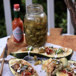 three venison stuffed zucchini boats on white cloth on wooden table with a jar of pickles and Tabasco sauce in background