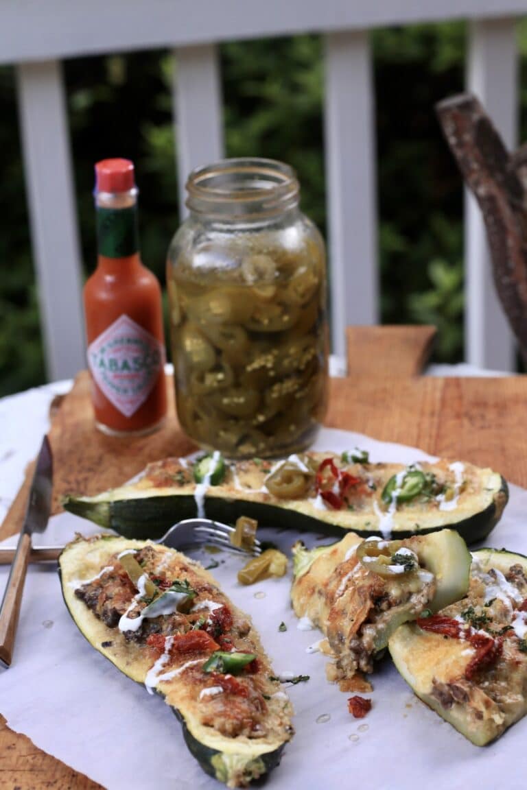 three venison stuffed zucchini boats on white cloth on wooden table with a jar of pickles and Tabasco sauce in background