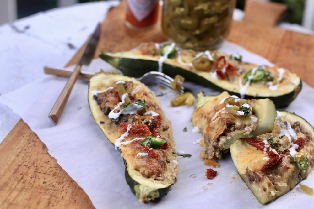 closeup of venison stuffed zucchini boats on white cloth on cutting board with fork, knife, and jar of pickes
