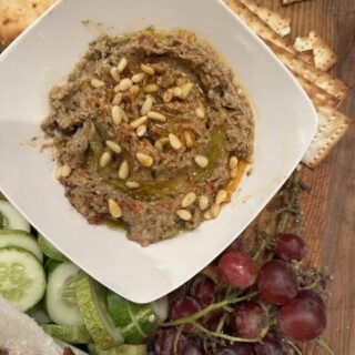 Stacy Lyn's baba ganoush, served in square white dish with pine nuts, surrounded by cucumbers, grapes, and pita chips on wood table