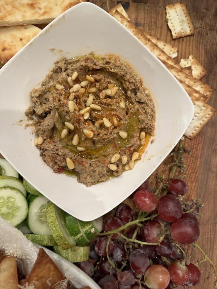 Stacy Lyn's baba ganoush, served in square white dish with pine nuts, surrounded by cucumbers, grapes, and pita chips on wood table