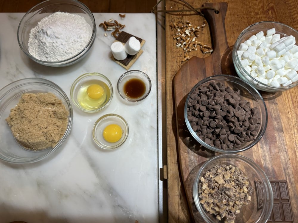 ingredients for s'mores cookie recipe in bowls on cutting board and kitchen counter: flour mixture, sugar, eggs, vanilla extract, crushed pretzel and Heath bar toffee, chocolate chips, marshmallows