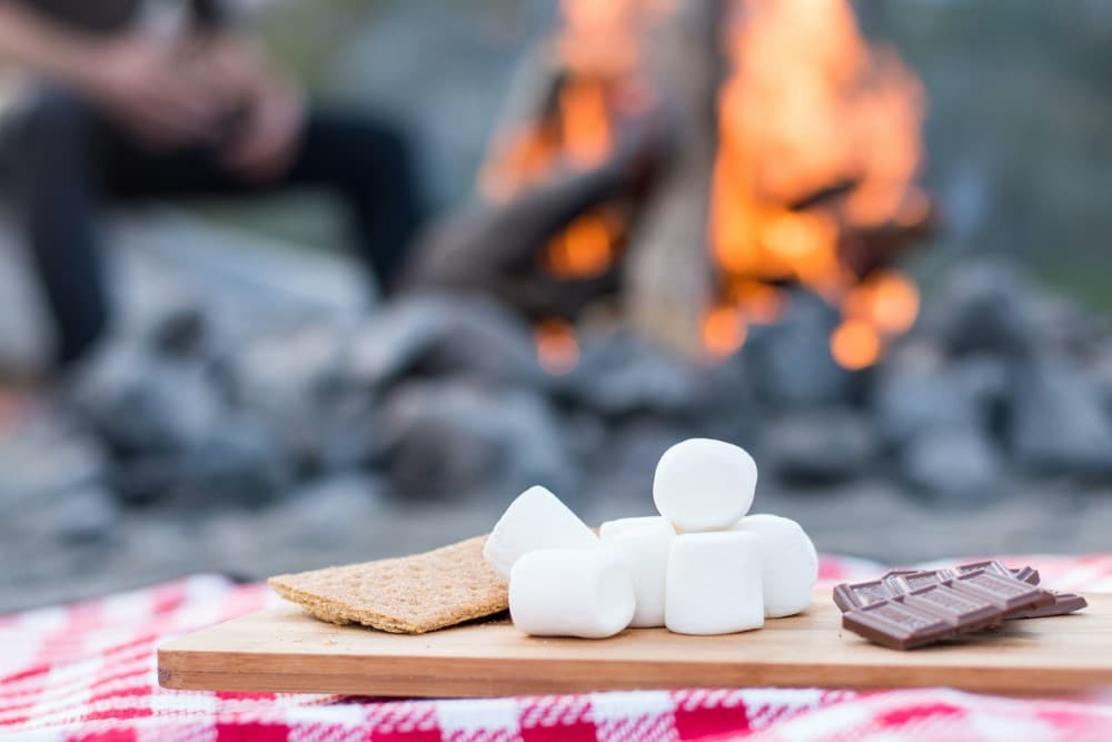 s'mores ingredients on red and white checkered tablecloth in front of a campfire: graham crackers, marshmallows, chocolate bars