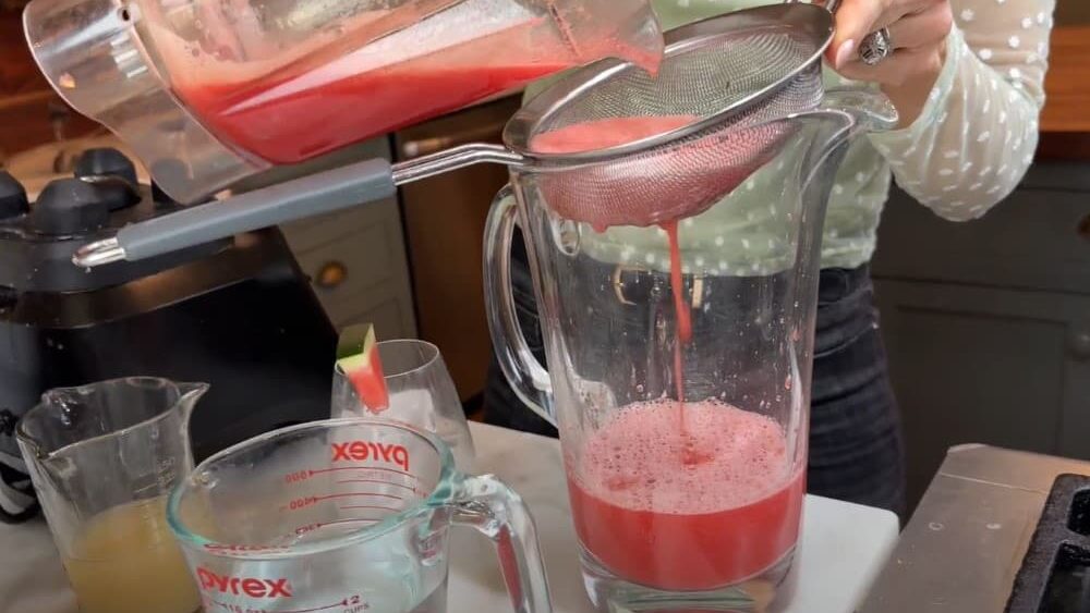 Stacy Lyn pouring blended watermelon through sieve into glass pitcher to strain out the pulp