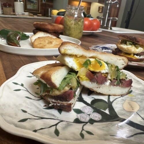 meat, egg, and green tomato relish sandwich sliced in half on leaf-design plate - two more plates and mason jar of green tomato relish, plate of fresh tomatoes in background on counter