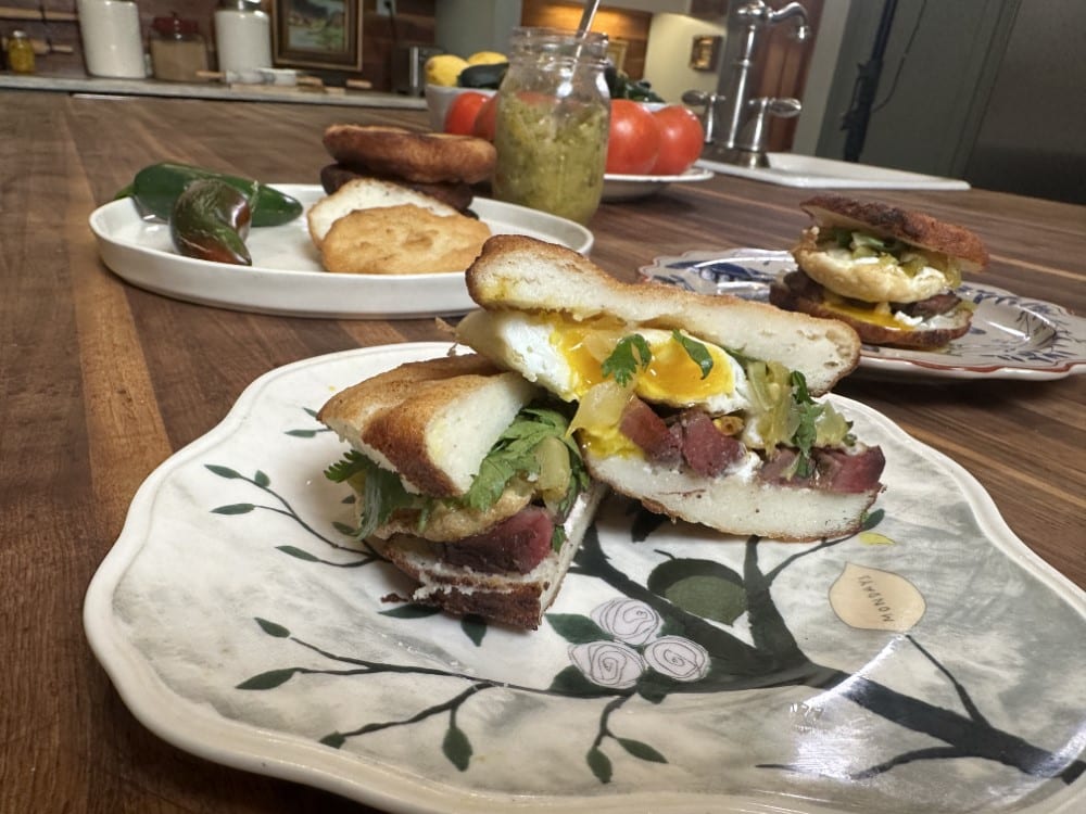 meat, egg, and green tomato relish sandwich sliced in half on leaf-design plate - two more plates and mason jar of green tomato relish, plate of fresh tomatoes in background on counter