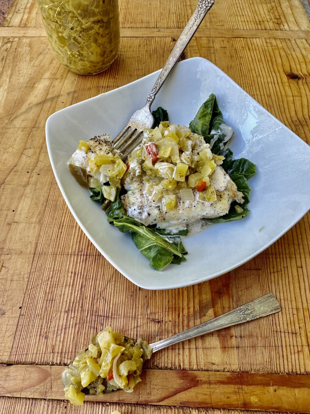 grilled halibut dish served with relish on bed of turnip greens on square white plate - fork is resting on plate, which sits on wood table with jar of green tomato relish in background