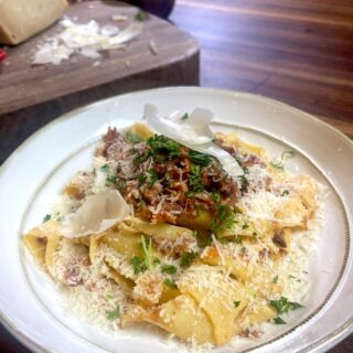 large white plate full of wild boar ragu over papardelle pasta with block of cheese on wooden cutting board in background
