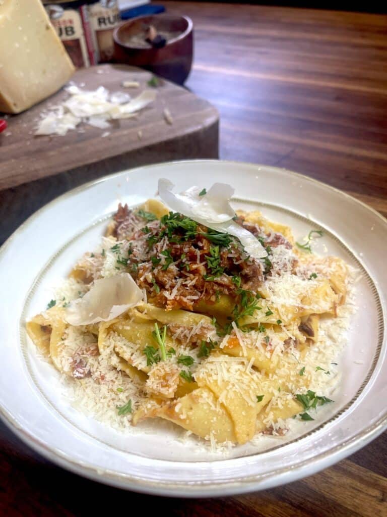 large white plate full of wild boar ragu over papardelle pasta with block of cheese on wooden cutting board in background