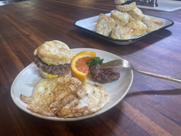 horizontal image of breakfast plate with fried eggs, sliced orange, bite of sausage on fork, and breakfast biscuit