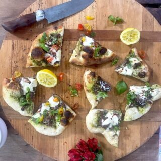 Wedges of turkey pizza with walnut basil pesto and burrata and parmesan cheese on circular wooden cutting board with lemon halves, flower, and knife