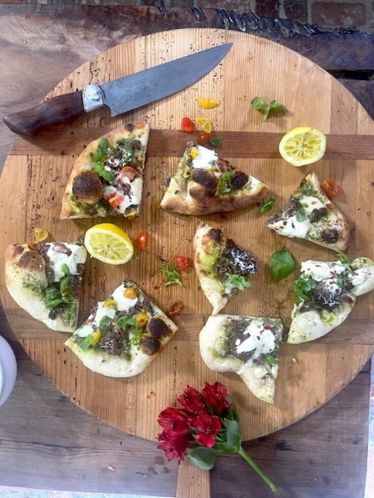 Wedges of turkey pizza with walnut basil pesto and burrata and parmesan cheese on circular wooden cutting board with lemon halves, flower, and knife