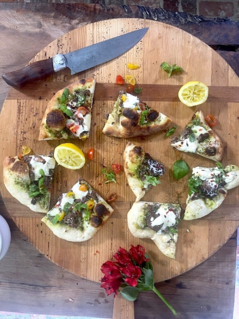 Wedges of turkey pizza with walnut basil pesto and burrata and parmesan cheese on circular wooden cutting board with lemon halves, flower, and knife
