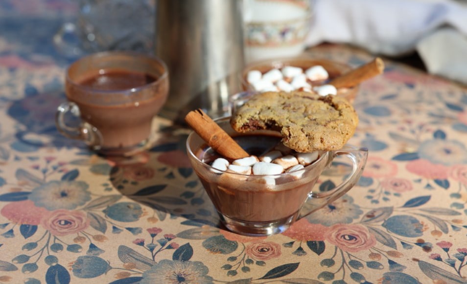 closeup of glass cups with homemade hot chocolate, marshmallows, cinnamon sticks on table with pretty cloth