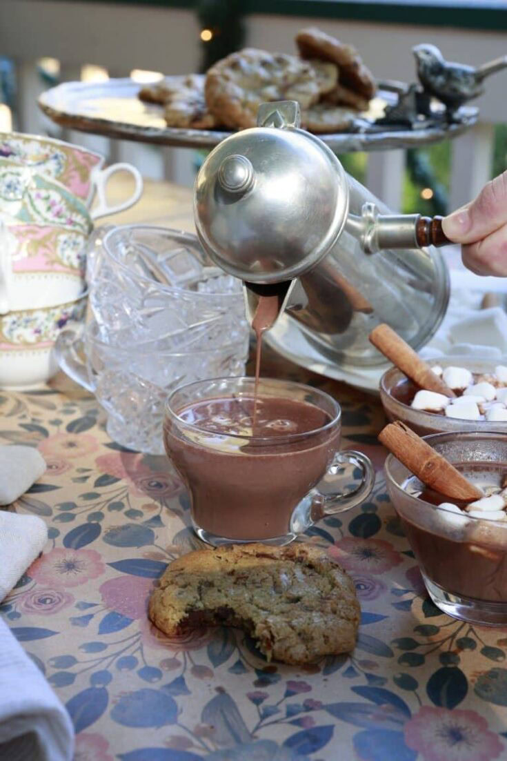 Stacy Lyn's hand pouring a cup of hot chocolate from a metal carafe - kitchen sink cookie with a bite out of it in foreground