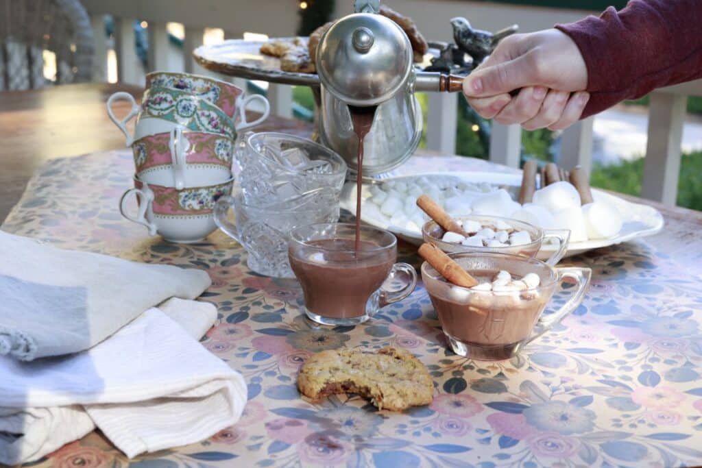 pouring hot cocoa from old-fashioned metal carafe into pretty glass cups on floral tablecloth - cinnamon and marshmallows in two cups - cookie in front - more ingredients in background