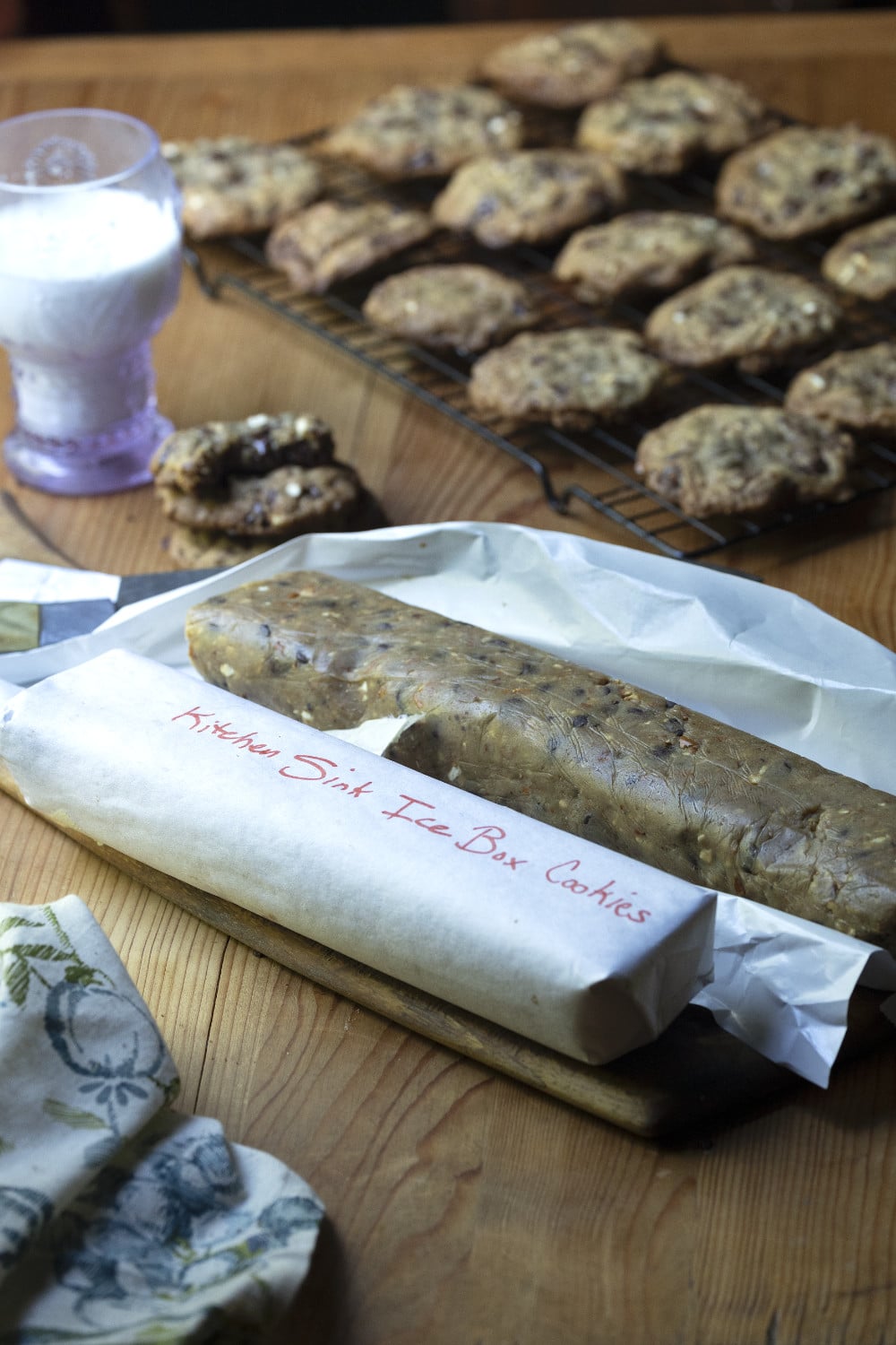kitchen sink cookies dough rolled in freezer paper to store for later baking - in background, a rack of freshly baked cookies and a glass of milk