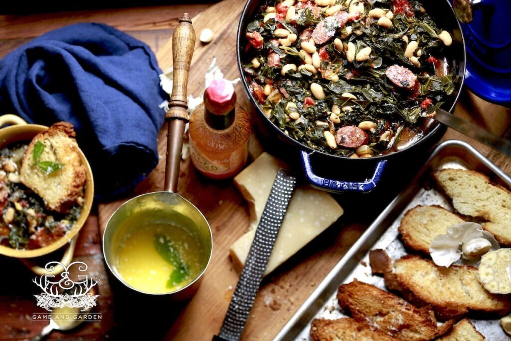 large ceramic lined Dutch oven of collards and bean soup on wooden table with baking tray of toasted bread, bowl of soup with slice of bread, blue kitchen towel, and cooking ingredients