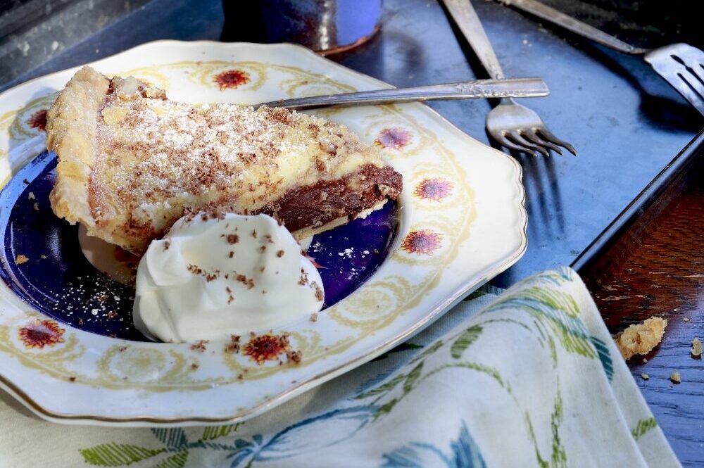 slice of black bottom pie on china plate with whipped cream - plate is on dark tray with cup of coffee, set on blue floral tablecloth