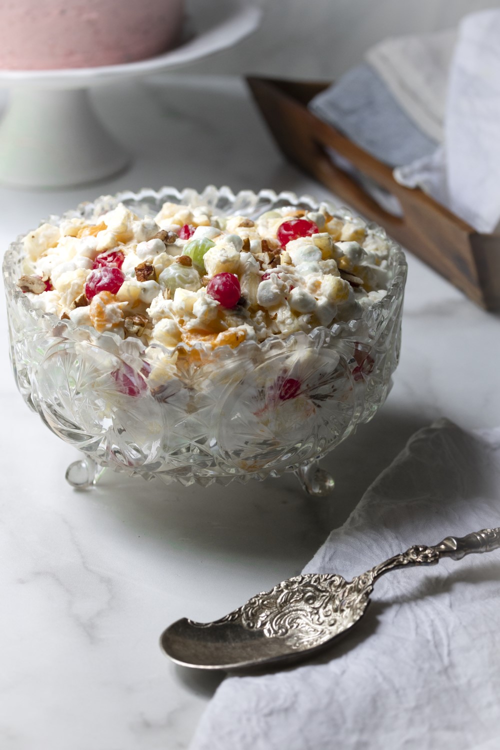 Ambrosia fruit salad dessert in cut crystal dish resting on white marble counter with antique silver spoon on white napkin