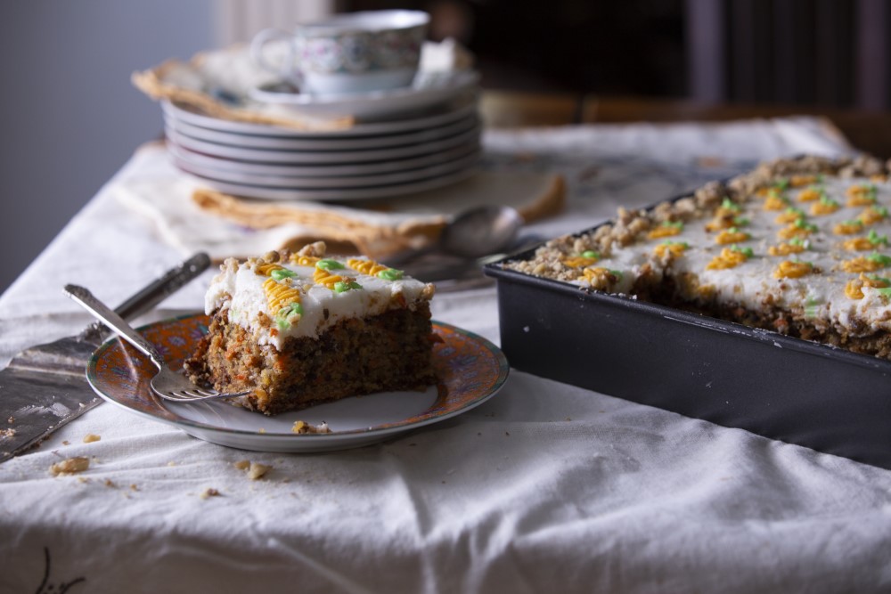 A aquare slice of carrot cake with cream cheese frosting next to sheet pan of cake - decorated with frosting carrots - and a stack of dessert plates with coffee cup