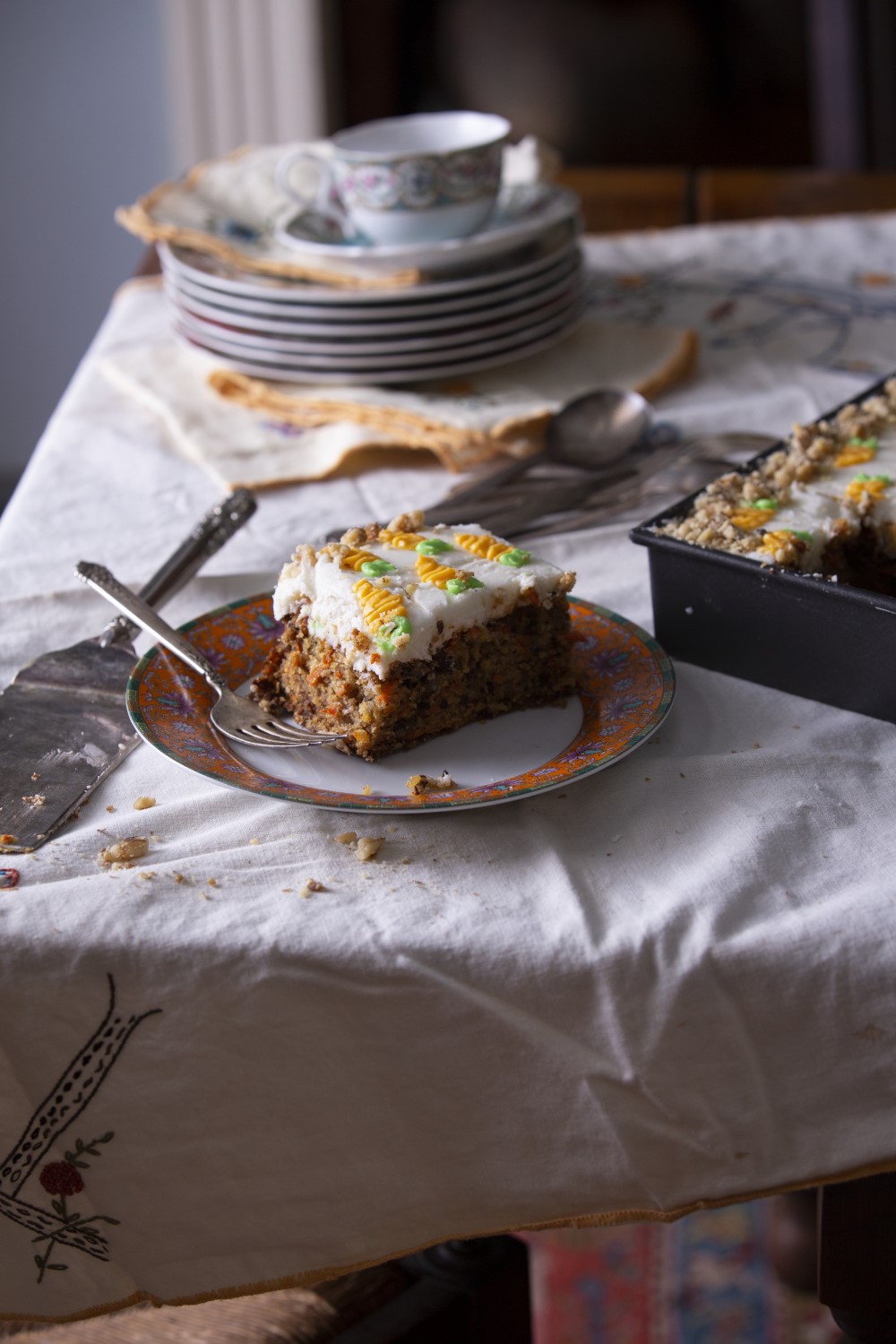 slice of carrot cake on floral dessert plate with fork on white tablecloth - cake knife to left and corner of sheet cake pan to right - stack of dessert plates with coffee cup in background
