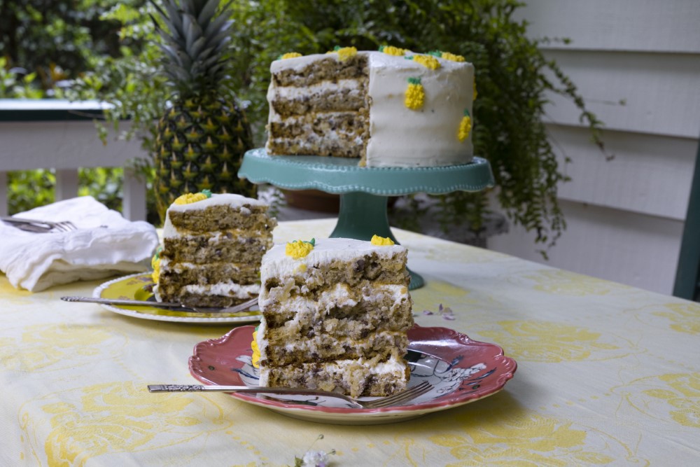 slices of 4-layer hummingbird cake and a fresh pineapple with full cake on cake plate on outdoor table