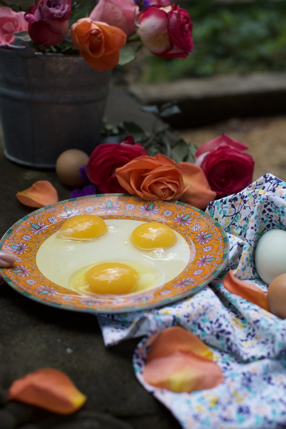 Floral pattern plate with three raw egg yolks,, nestled on bed of roses and napkin with tin bucket of roses in background