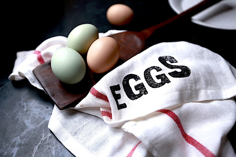 four fresh eggs, two brown and two white, on wooden spatula atop dish towel labeled EGGS