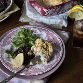 serving of poppy seed chicken casserole on China plate with salad - red floral casserole dish and glass of iced tea in background