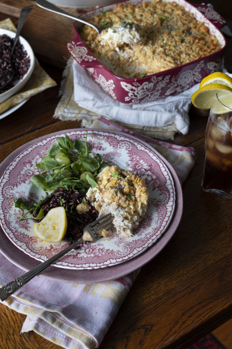 serving of poppy seed chicken casserole on China plate with salad - red floral casserole dish and glass of iced tea in background