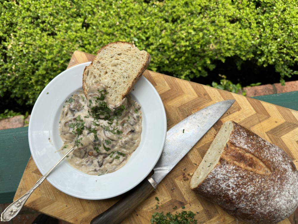 cream of mushroom soup in white bowl with slice of bread balanced on rim, sliced loaf of bread and knife on cutting board