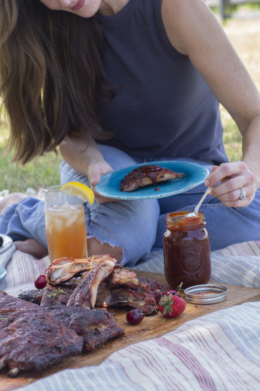 Stacy Lyn sitting on picnic blanket outdoors in blue tank top and torn jeans, smiling while spooning BBQ sauce from a mason jar onto a plate of grilled BBQ ribs