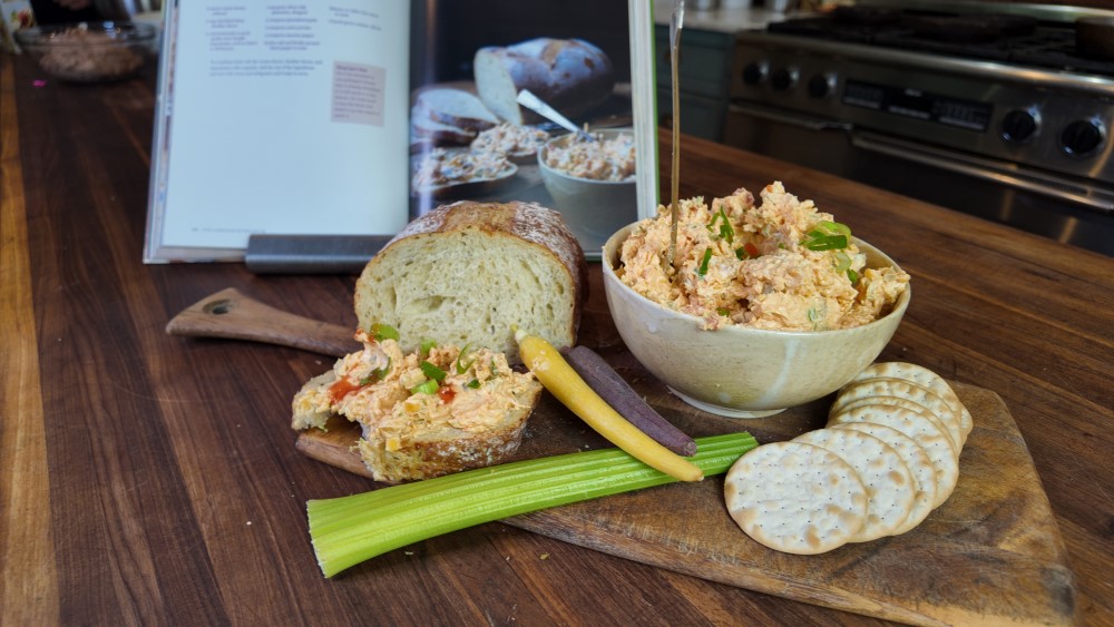 heaping bowl of pimento cheese spread on wooden cutting board with celery, pimento cheese sandwich, and crackers - Stacy Lyn's Love Language of the South cookbook open to recipe in background