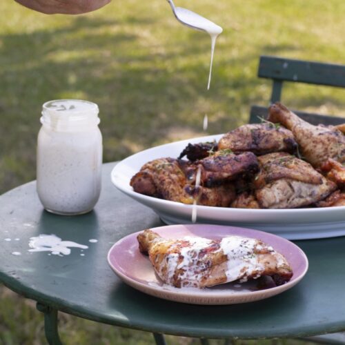 smoked chicken on serving platter on outdoor table with plated serving in front, hand drizzling white sauce onto chicken