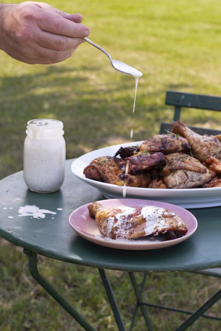 smoked chicken on serving platter on outdoor table with plated serving in front, hand drizzling white sauce onto chicken