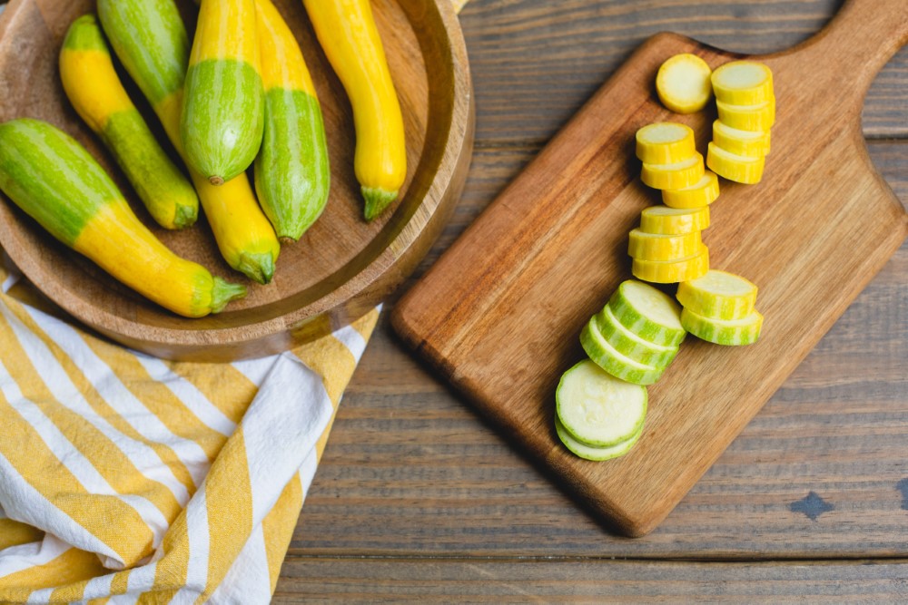 Top View of Fresh Zephyr Squash Sliced on a Wooden Cutting Board; Whole Squash in a Wooden Bowl on a Wooden Table in Background