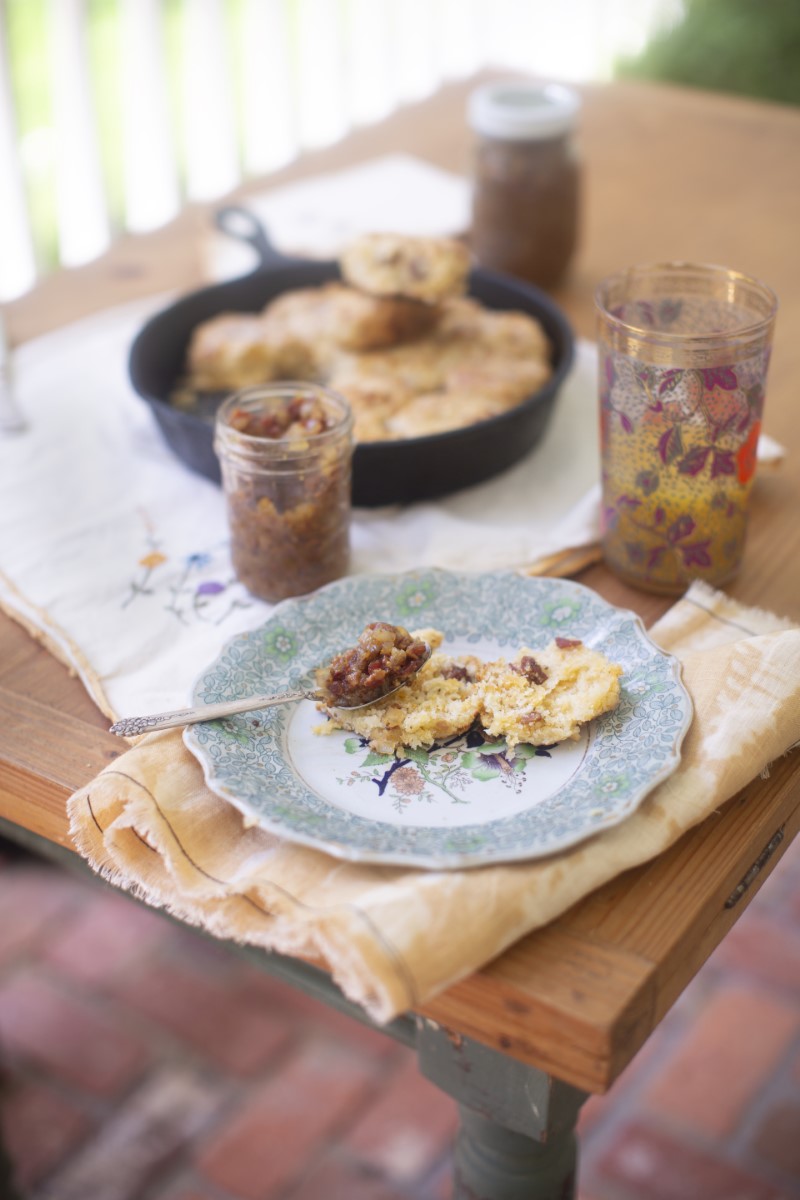 Halved buttermilk cheddar biscuit on blue floral china breakfast plate with spoonful of bacon jam - folded cloth napkin and drinking glass - skillet full of freshly baked biscuits on table in background