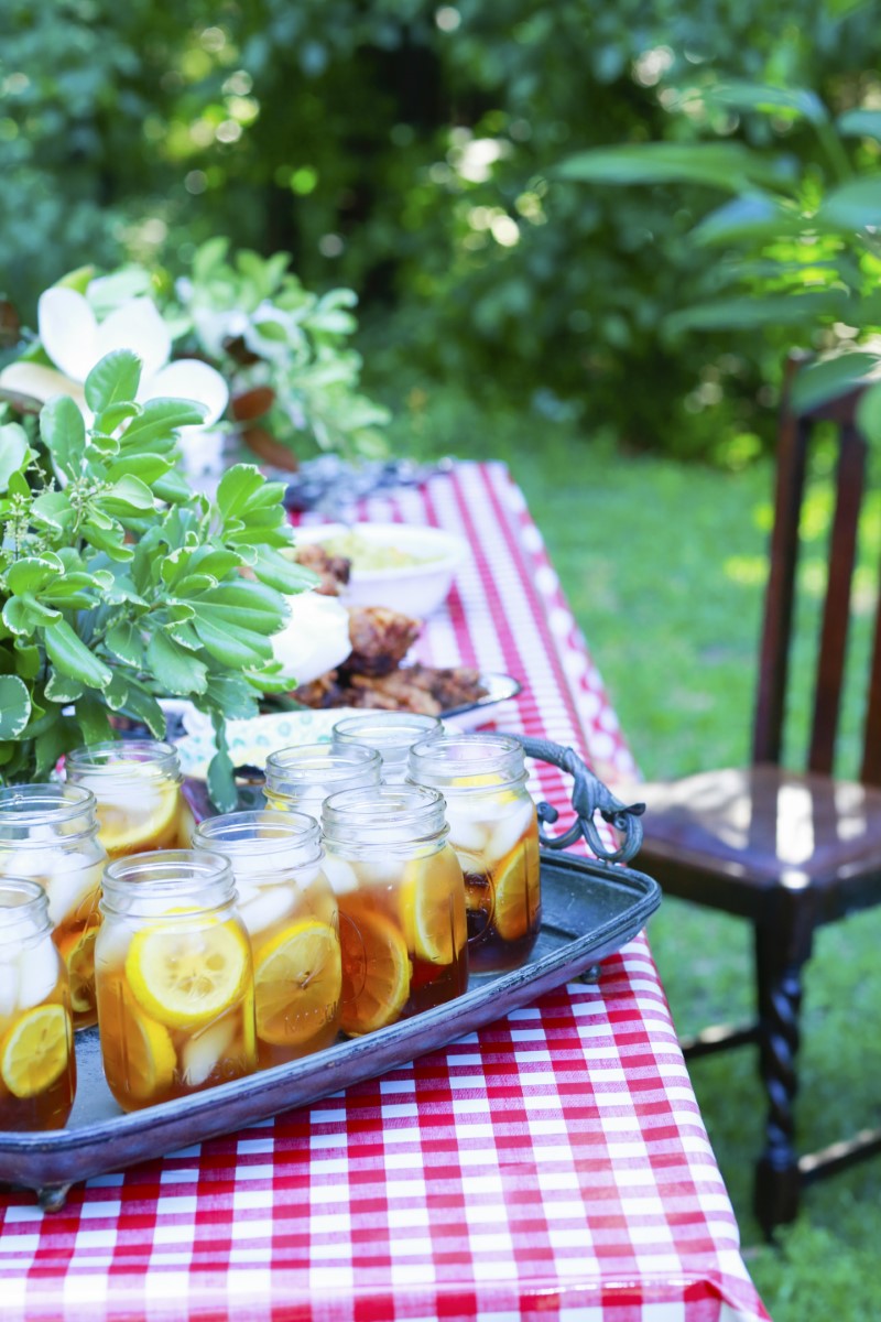 sweet tea served in mason jars with ice on a red checkered cloth outdoors next to a fried chicken meal