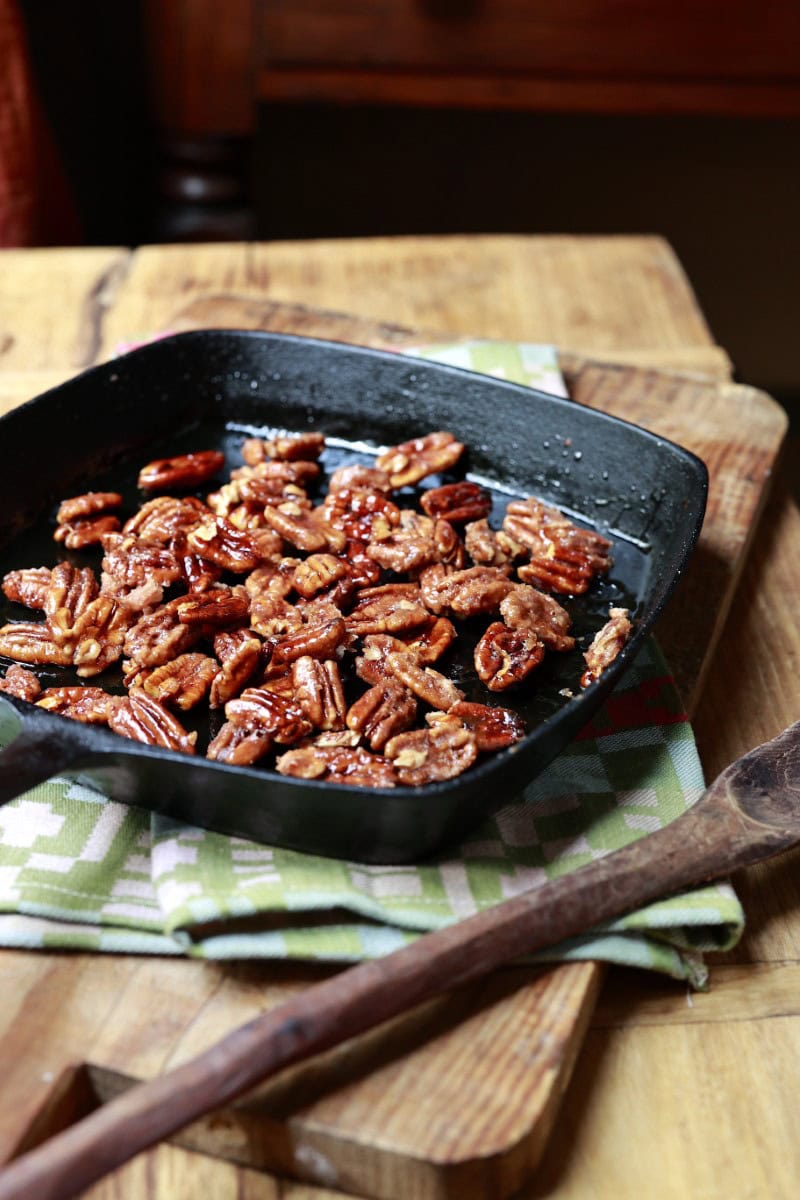 candied pecans in cast-iron skillet resting on green dishcloth alongside wooden spoon on chopping block table