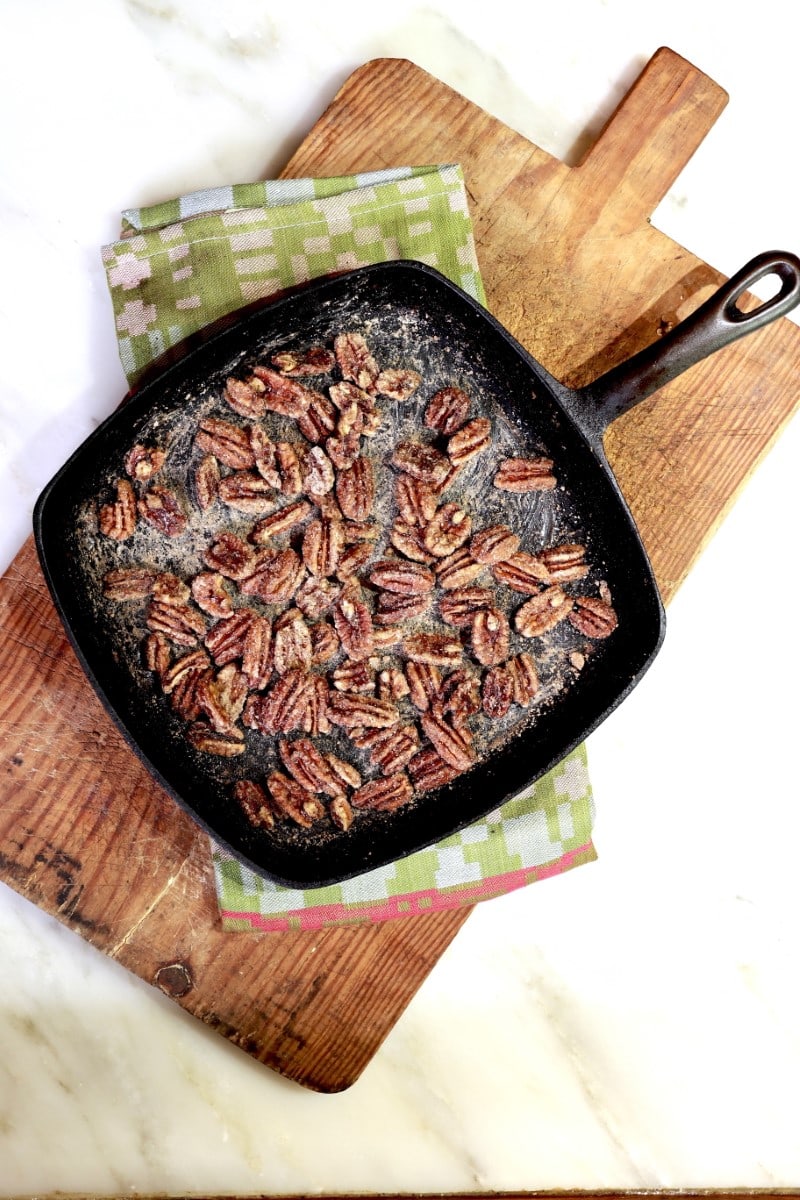 vertical shot of candied pecans in square skillet resting on green dishcloth atop wooden cutting board