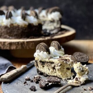Oreo cheesecake slice on rustic stone plate with fork and knife and with cheesecake in background with slice out of it