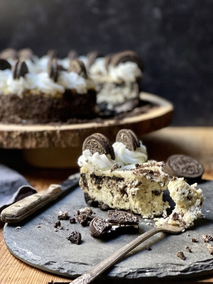 Oreo cheesecake slice on rustic stone plate with fork and knife and with cheesecake in background with slice out of it