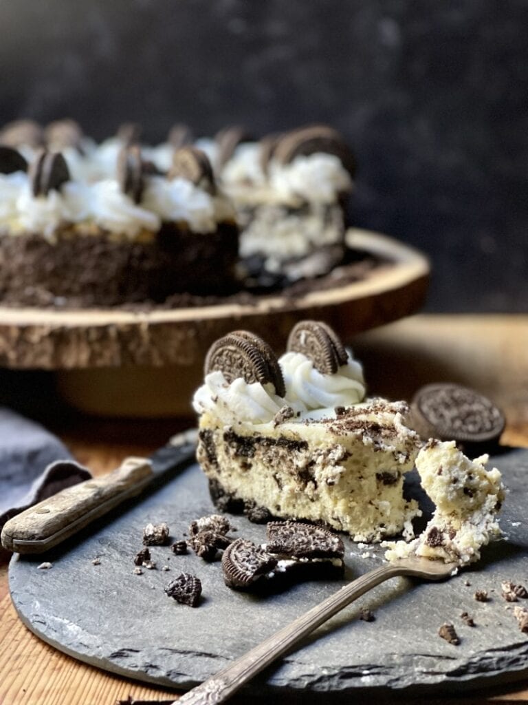 Oreo cheesecake slice on rustic stone plate with fork and knife and with cheesecake in background with slice out of it