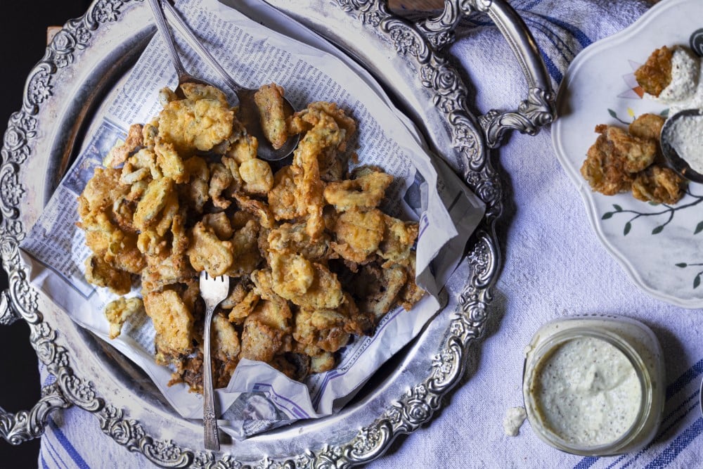 fried pickles on newspaper-lined silver platter with jar of creamy sauce and serving plate on side
