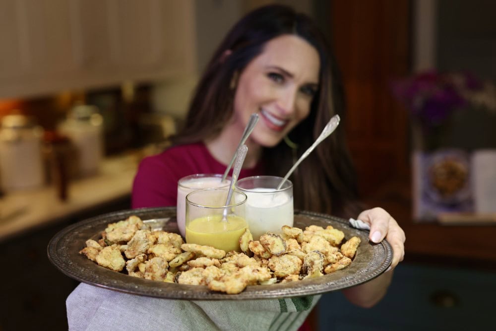 Stacy Lyn Harris in her kitchen holding out platter of fried pickles with several dipping sauces