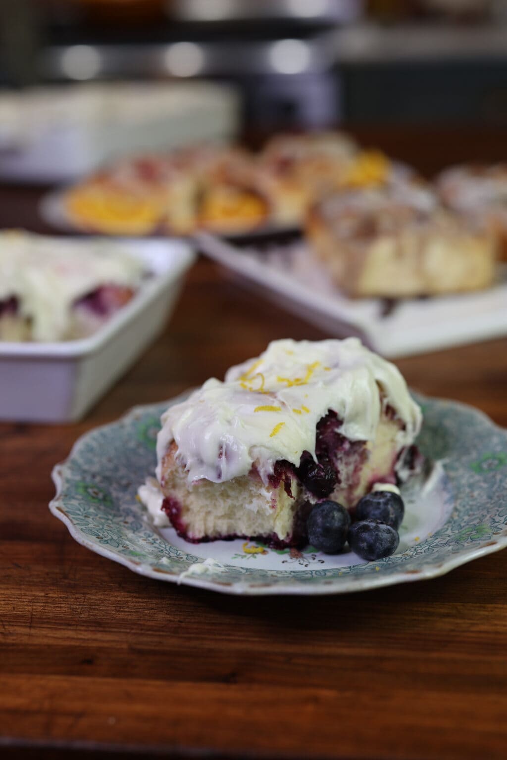 frosted blueberry roll on plate in front of pans full of cinnamon rolls