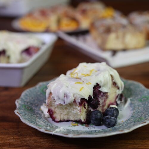 frosted blueberry roll on plate in front of pans full of cinnamon rolls