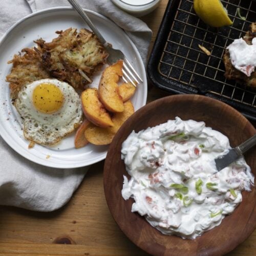 white plate with crispy potato latkes alongside over easy egg and peach slices - wood bowl with sour cream lox sauce - more latkes on cookie sheet in back